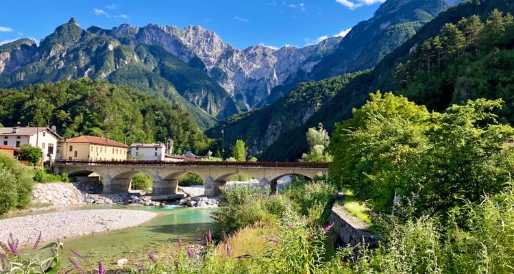 Puente de piedra que cruza un río turquesa rodeado de montañas boscosas empinadas y cielos azules despejados.