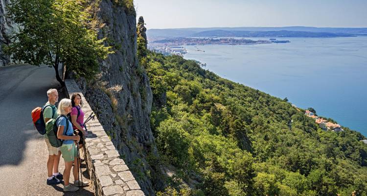 Tres caminantes se encuentran en un sendero del acantilado contemplando el Golfo de Trieste y su puerto.