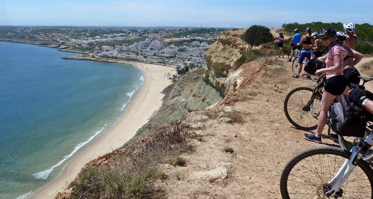 Groupe de cyclistes en pause sur un sentier de falaise côtière surplombant une plage de sable et une ville balnéaire.