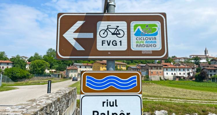 Richtungsschild mit Fahrradroutensymbolen und malerischem Dorf im Hintergrund.