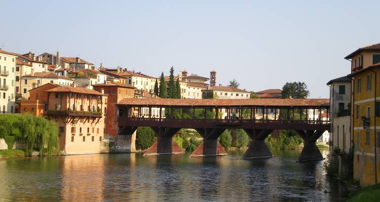 Historic wooden covered bridge spans the Brenta River beside pastel Italian buildings.
