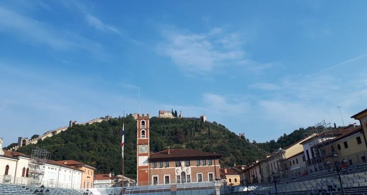 Walled hillside castle overlooks open square and medieval town under a blue sky.