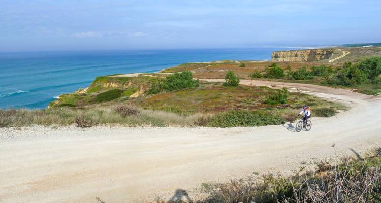 Cycliste sur un sentier de terre surplombant la mer avec des falaises et de la végétation.