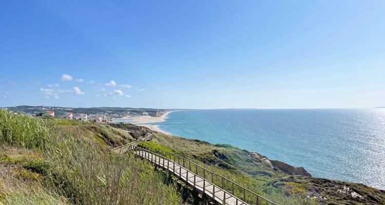Ein hölzerner Steg schlängelt sich über grasbewachsene Klippen zu einem langen Sandstrand und dem azurblauen Meereshorizont.