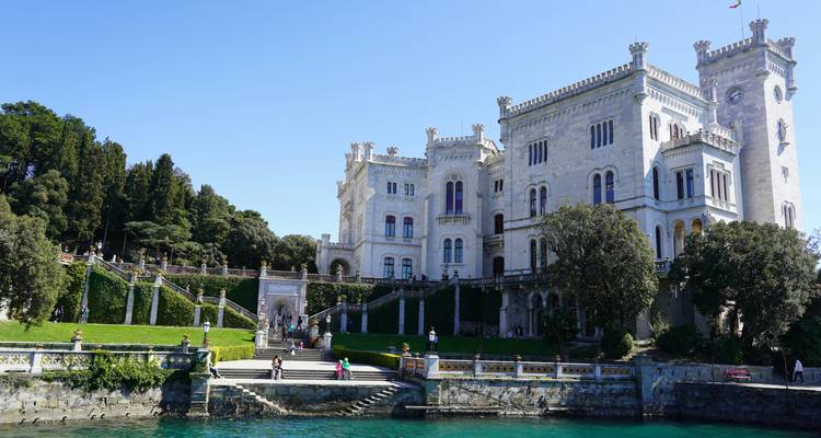 Großes historisches schlossähnliches Gebäude am Wasser mit Menschen auf Treppen.