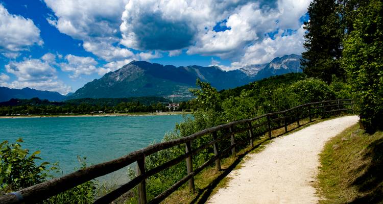 Weg entlang eines Seeufers mit Bergen im Hintergrund.