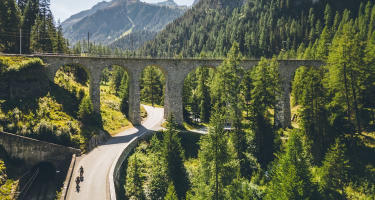 Une vue panoramique d'un viaduc en pierre avec des cyclistes en contrebas, entouré d'une végétation luxuriante et d'un arrière-plan montagneux.