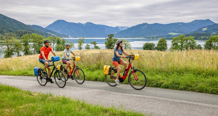 Drei Radfahrer fahren auf einer Landstraße oberhalb eines breiten Sees, der von Bergen umgeben ist.