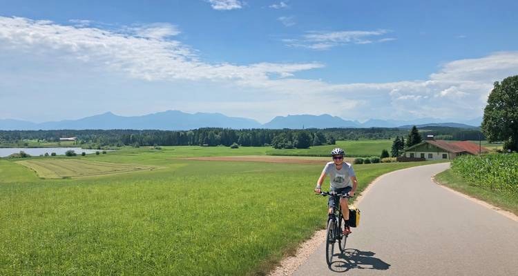 Eenzame fietser die peddelend over een rustige landweg rijdt door open groene velden met bergen aan de horizon.