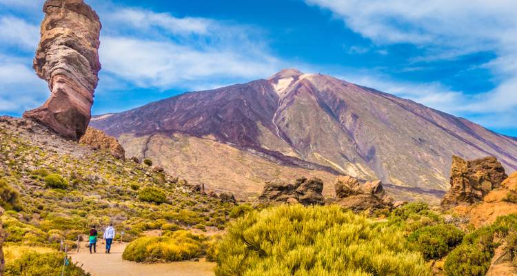 Two hikers walking in a volcanic landscape with a prominent rock formation.