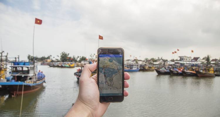 Een hand die een telefoon vasthoudt met een kaart, boten op de achtergrond.