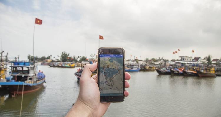 Main tenant un smartphone affichant une carte devant un port rempli de bateaux de pêche vietnamiens et de drapeaux.
