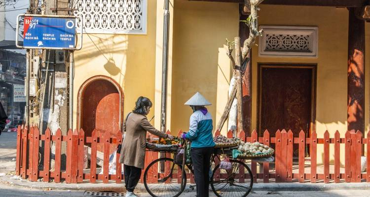 Deux femmes, l'une poussant un chariot à vélo de marchandises, conversent devant une maison jaune dans une rue tranquille vietnamienne.