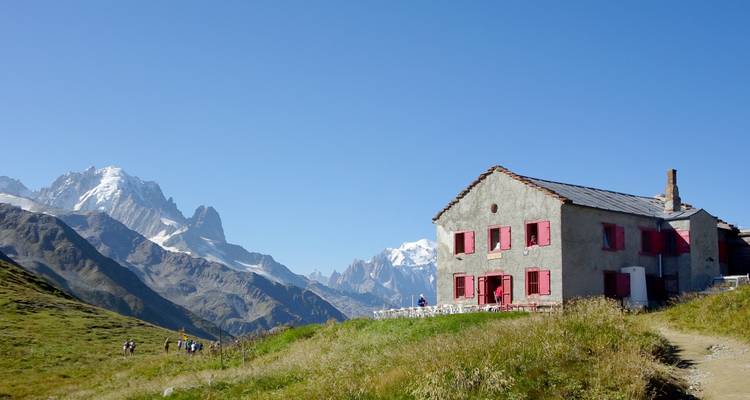 A rustic mountain refuge with bright red shutters overlooks the snowy peaks of the Mont Blanc massif while hikers approach along a grassy ridge.