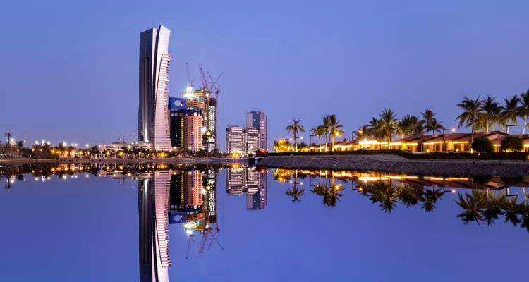 Wolkenkratzer und Palmen spiegeln sich in einem ruhigen Meer am Abend.