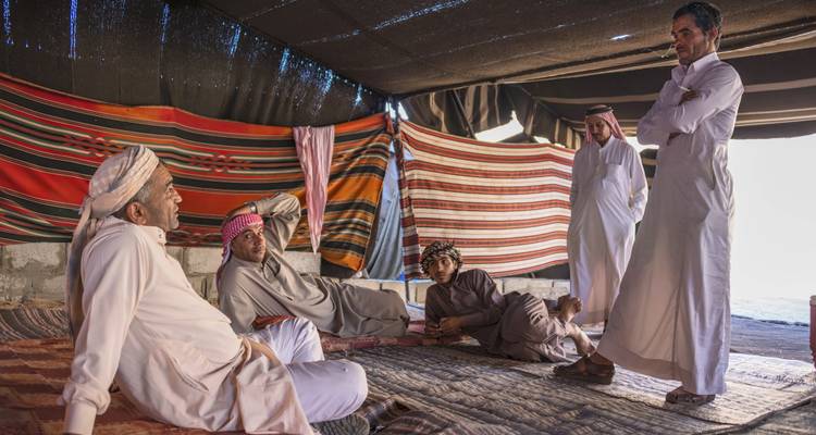 Bedouin men in traditional dress relax and converse inside a colorful tent lined with striped rugs.