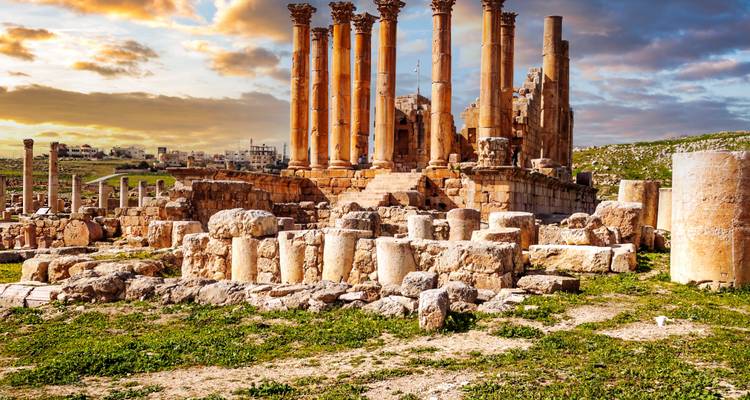 Gut erhaltene römische Säulen und Ruinen von Jerash, beleuchtet durch goldenen Sonnenuntergang mit Stadt im Hintergrund.