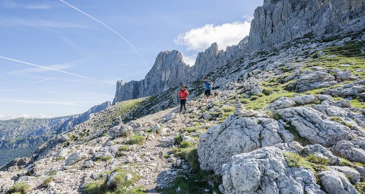 Hikers walking along a rocky trail with stunning mountain cliffs in the background.