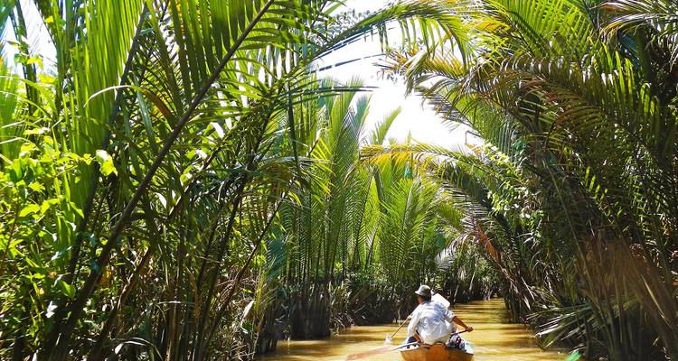 Man paddling a small boat along a waterway surrounded by dense vegetation.