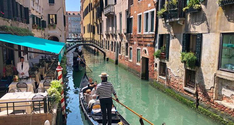 Ein Gondoliere navigiert durch einen schmalen venezianischen Kanal vorbei an Cafés am Wasser und blumengeschmückten Balkonen.