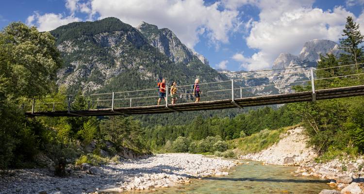 Des personnes marchant sur un pont suspendu au-dessus d'une rivière avec des montagnes en arrière-plan.