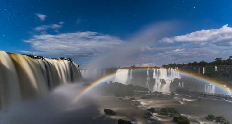 Spektakulärer Nachtblick auf die Iguazú-Wasserfälle mit einem nebelumhüllten Regenbogen unter sternenklarem Himmel.