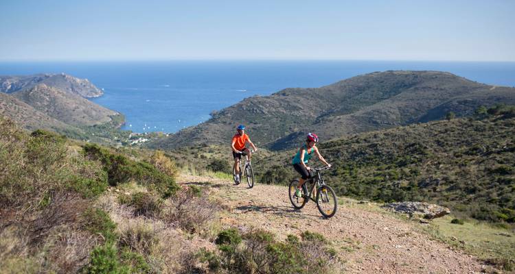 Cyclists ride on a trail near the coast with hills and sea in view.