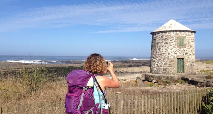 A person with a backpack taking a photo of a stone building by the sea.