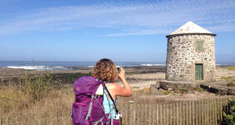A person with a backpack taking a photo of a stone windmill by the sea.