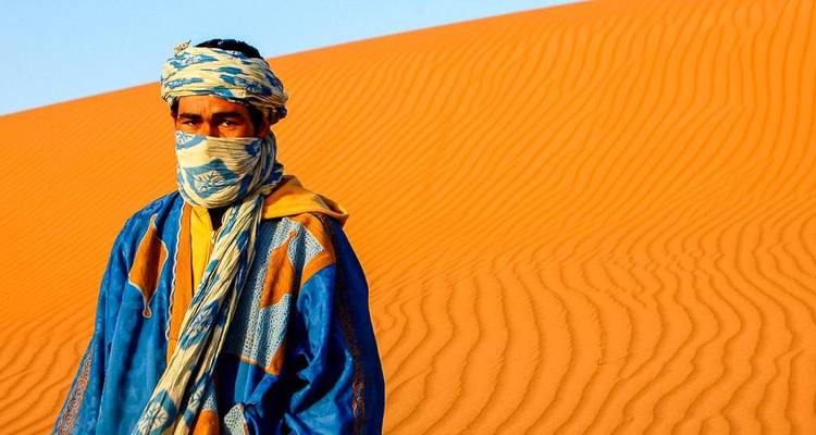 A man in traditional attire standing in front of a vast sand dune.