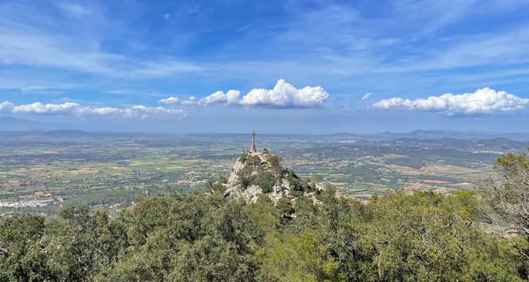 Cumbre montañosa coronada con una gran cruz que domina extensas llanuras y cordilleras distantes bajo un cielo vívido.