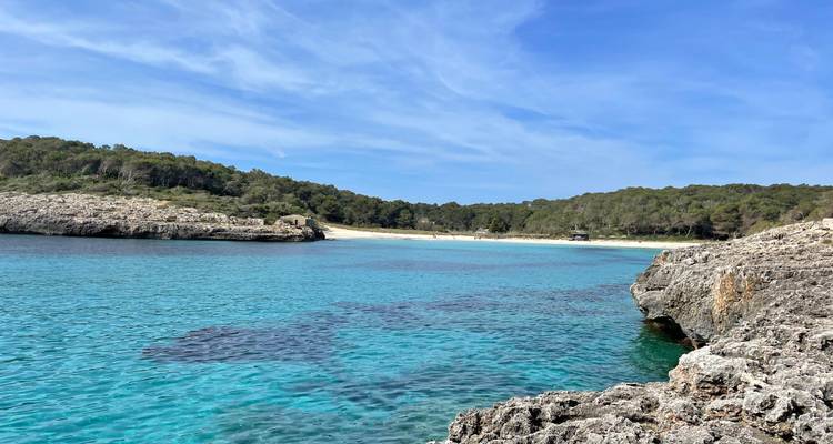 Bahía turquesa cristalina con playa de arena respaldada por promontorios boscosos, vista desde la orilla rocosa.