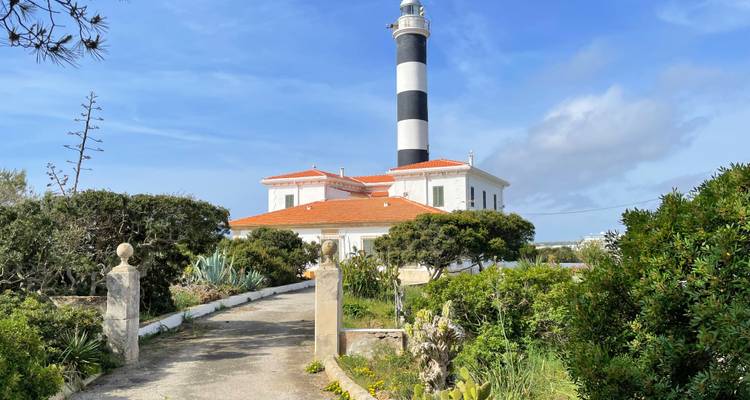 Faro de rayas blancas y negras con casa del farero de techo rojo rodeado de matorral mediterráneo.