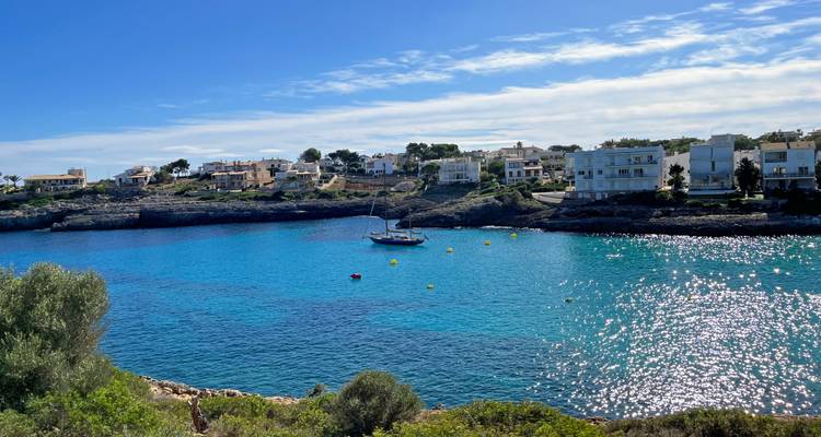 Bahía turquesa tranquila salpicada de boyas de amarre y un velero, bordeada por villas costeras blancas.