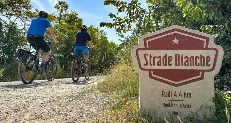 Zwei Radfahrer fahren auf der Schotter-Strade Bianche neben einem Straßenmarkierungsstein unter sonnigem toskanischem Himmel.