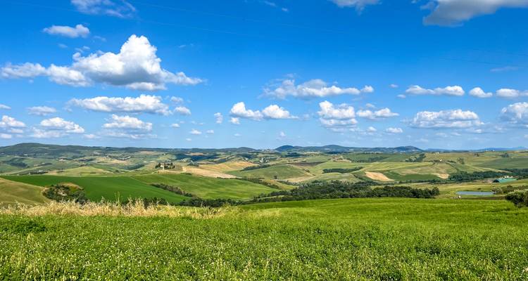 Weite, sanft geschwungene toskanische Landschaft mit flickenartigen Feldern und fernen Hügeln unter strahlend blauem Himmel.