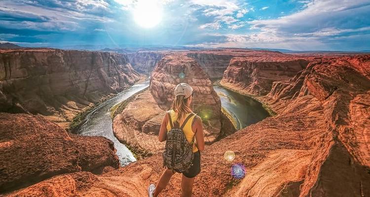 Person standing on a cliff overlooking a river bend and canyon under a sunny sky.