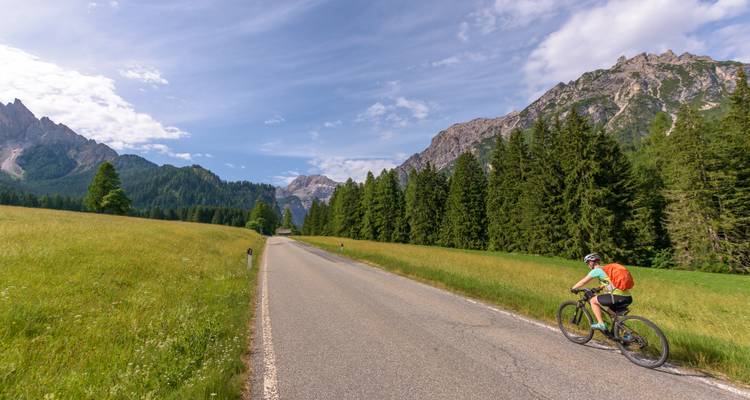 Cycliste sur une route pittoresque entourée de montagnes et de forêt sous un ciel lumineux.