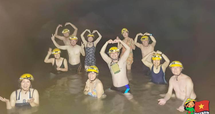 Un grand groupe de touristes se tient dans l'eau d'une grotte jusqu'à la taille en portant des casques ; autocollant cartoon du Vietnam visible.