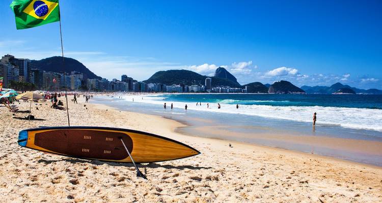 Scène de plage avec planche de surf, drapeau brésilien et personnes sur le sable.