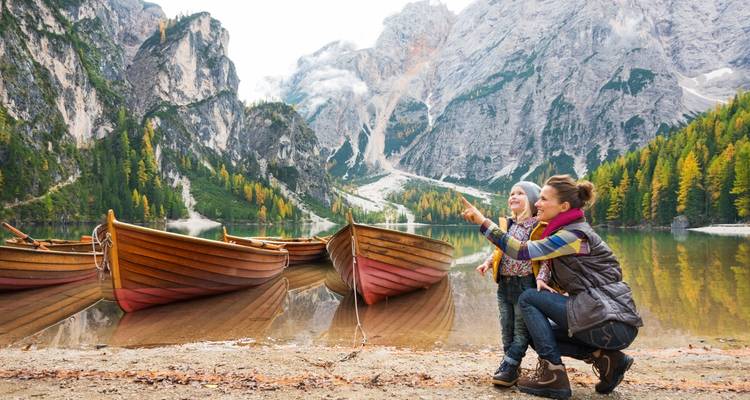 Mother and child near boats on a scenic lake with mountains.