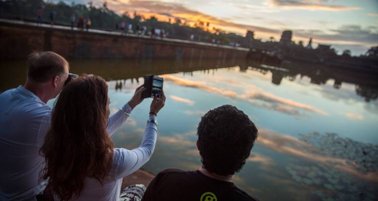 Los viajeros se sientan en un malecón de piedra fotografiando los reflejos del atardecer de las ruinas del templo al otro lado del foso.