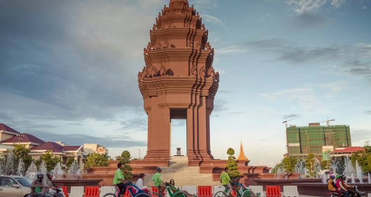 El tráfico nocturno de ciclistas y tuk-tuks pasa por el Monumento de la Independencia de piedra roja de Phnom Penh bajo un cielo pastel.