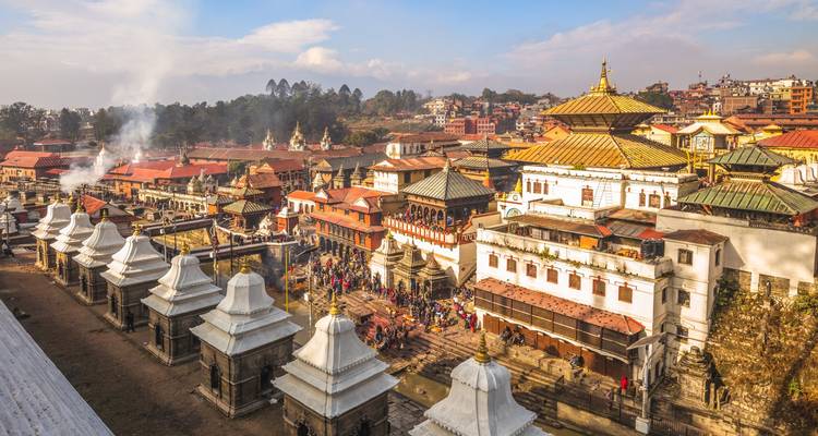 Vue aérienne d'un complexe de temples avec une végétation luxuriante, fumée visible des rituels de crémation.