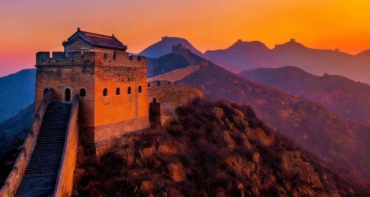 Dramatic sunset over the Great Wall of China, with shadowy silhouettes of mountains.