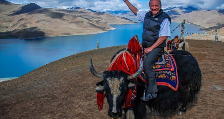 Man smiling while riding a yak with scenic mountain lake backdrop.