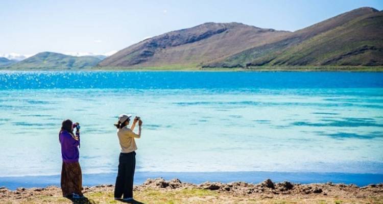 Two people taking photos by a scenic blue lake with hills in the background.