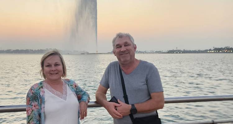 Deux personnes posant devant une grande fontaine avec un paysage urbain au coucher du soleil.