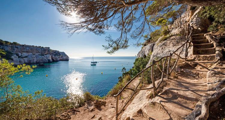 Seaside cliff path with a view of a sailboat and the sun reflecting on the water.