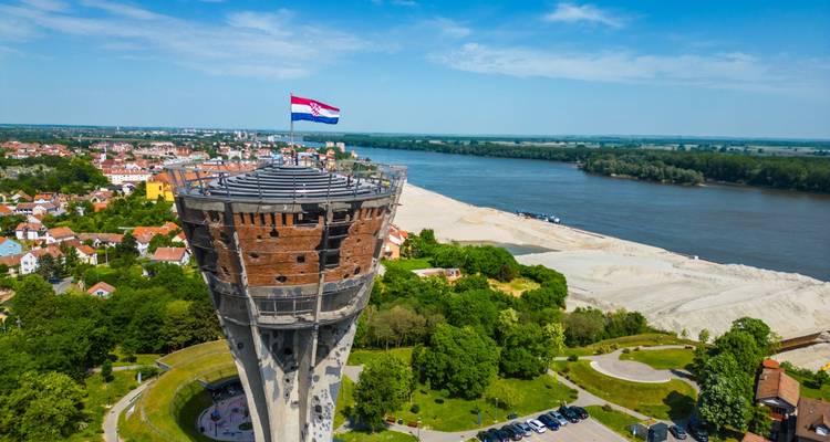 Vista aérea de una torre de agua con una bandera croata, rodeada de vegetación y un río.